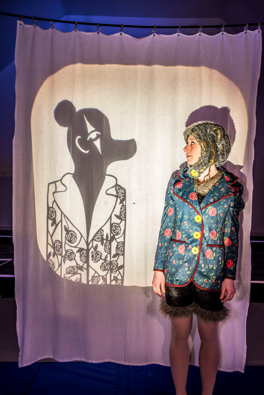 Picture of a woman in a bear hat and flowered jacket staring at a shadow puppet of a bear in a flowered jacket