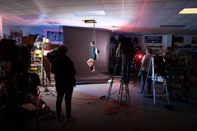 Photo of people standing around at a photo shoot as a white woman sits on a trapeze.