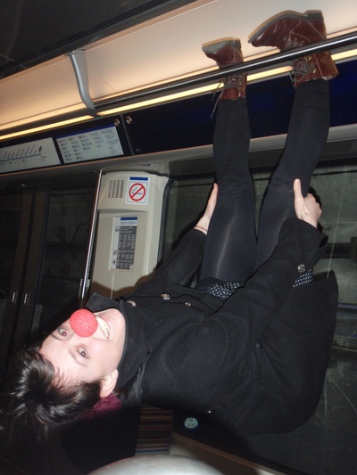 Photo of a woman with a clown nose hanging by her feet to the handrail of a Paris Metro car. 