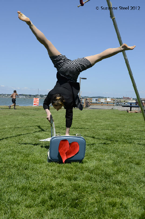Photo of a woman doing a handstand on a tiny chair. In front of her is a tiny suitcase with a paper broken heart put back together.