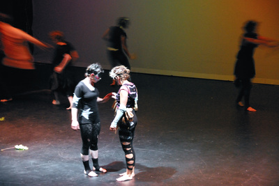 Photo of two white women in costumes touching hands onstage. Blurred people run in the background behind them.